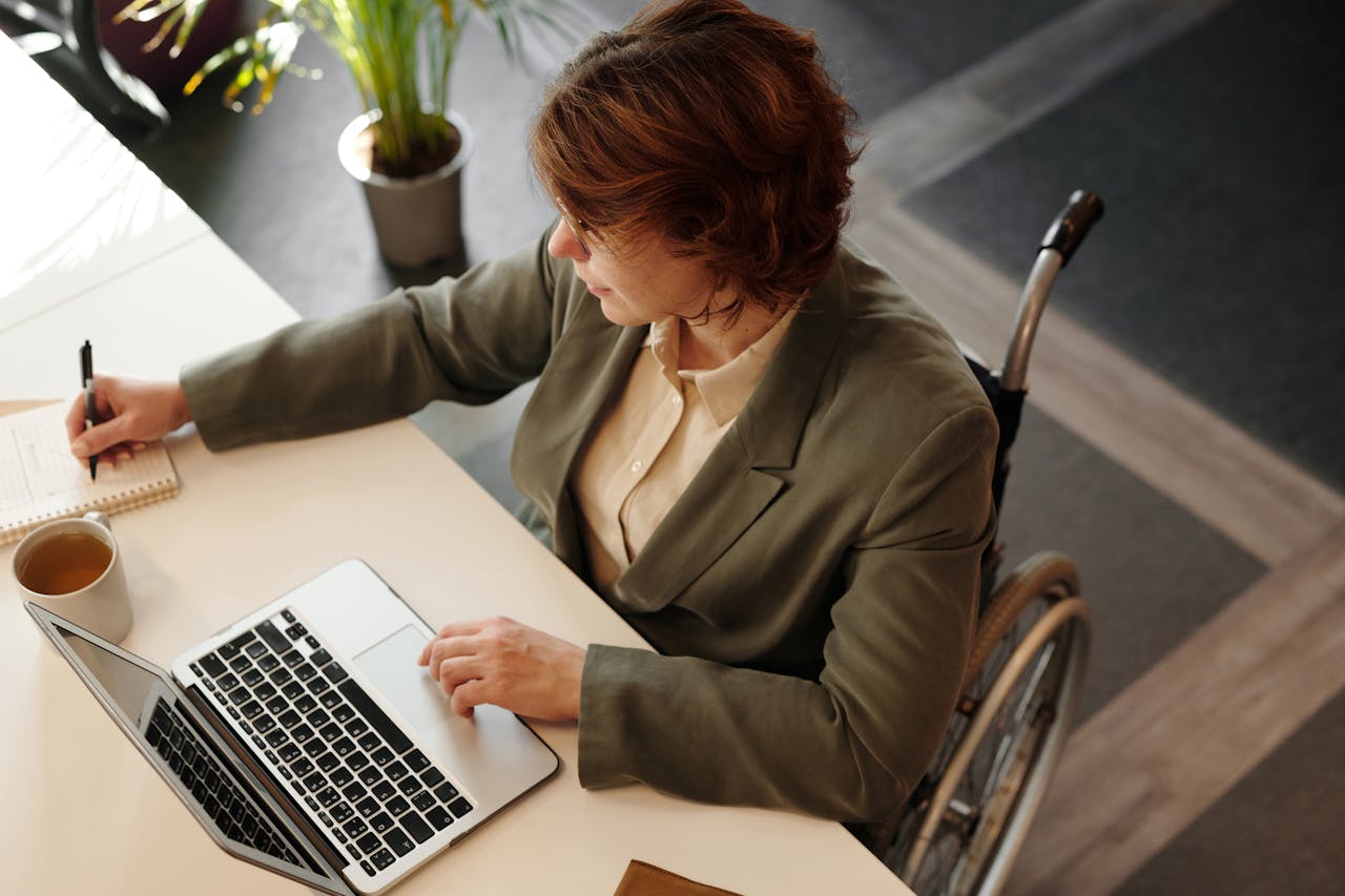 Top view of a businesswoman in a wheelchair working at her desk with a laptop and notebook.